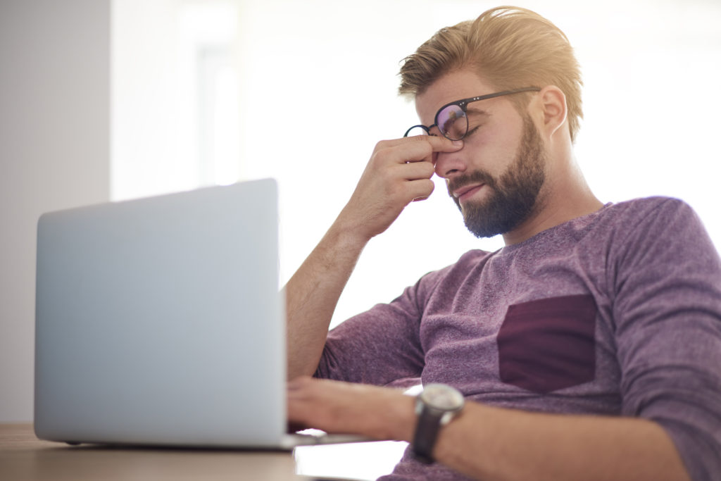 A msn wearing glasses sitting infront of a computer rubbing his eyes, he may be suffering from Dysfunctional Lens Syndrome