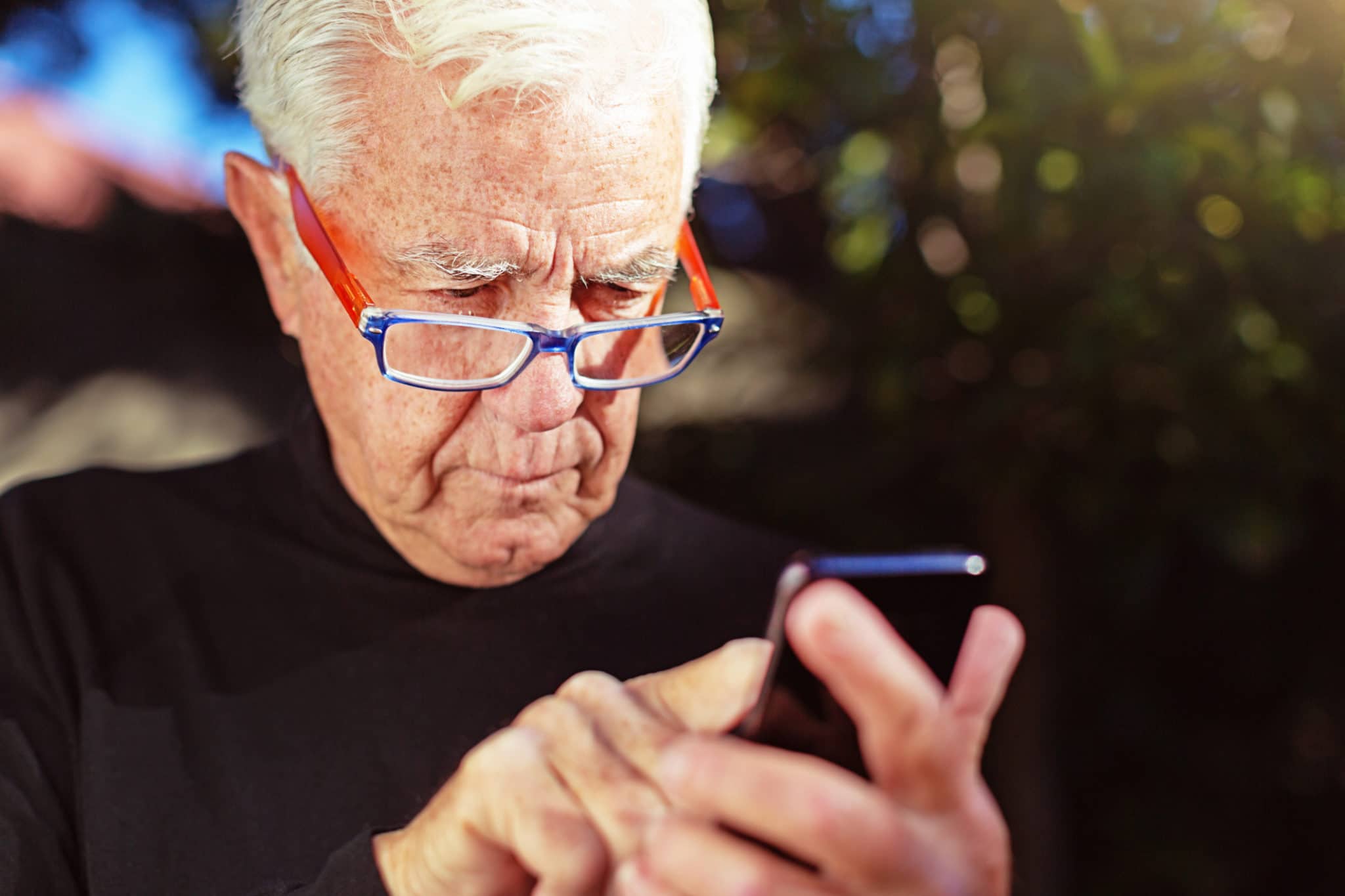 A senior man wearing eyeglasses peers down at his smart phone as he taps out a text.