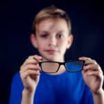 A portrait of a boy holds eyeglasses in hands in front of dark background. Children and healthcare concept
