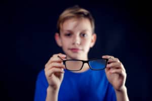 A portrait of a boy holds eyeglasses in hands in front of dark background. Children and healthcare concept